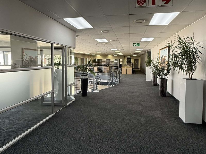 A spacious, well-maintained industrial office hallway featuring carpeted floors, suspended ceilings with recessed lighting, and glass partition walls overlooking the lower level.