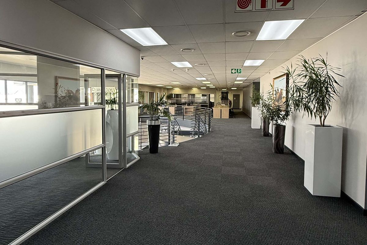 A spacious, well-maintained industrial office hallway featuring carpeted floors, suspended ceilings with recessed lighting, and glass partition walls overlooking the lower level.