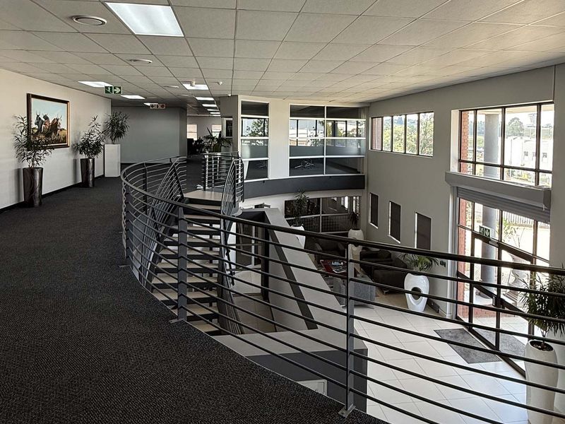 Spacious industrial office mezzanine level featuring a metal balustrade, carpeted walkway, and large windows overlooking the ground floor entrance area.