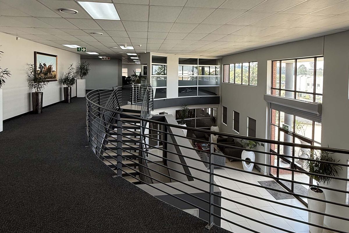 Spacious industrial office mezzanine level featuring a metal balustrade, carpeted walkway, and large windows overlooking the ground floor entrance area.