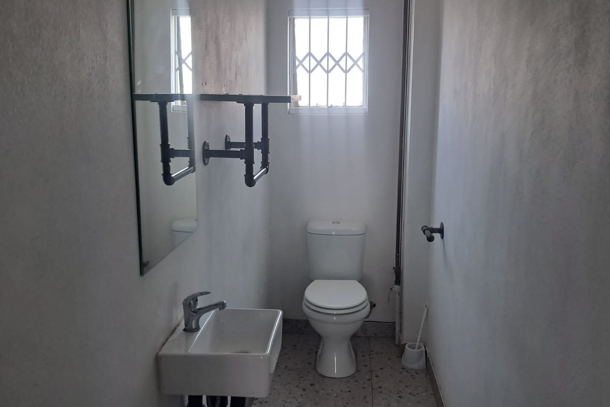 A compact industrial-style bathroom featuring a wall-mounted basin, toilet, and security-barred window, requiring minor cosmetic updates to the wall finishes.