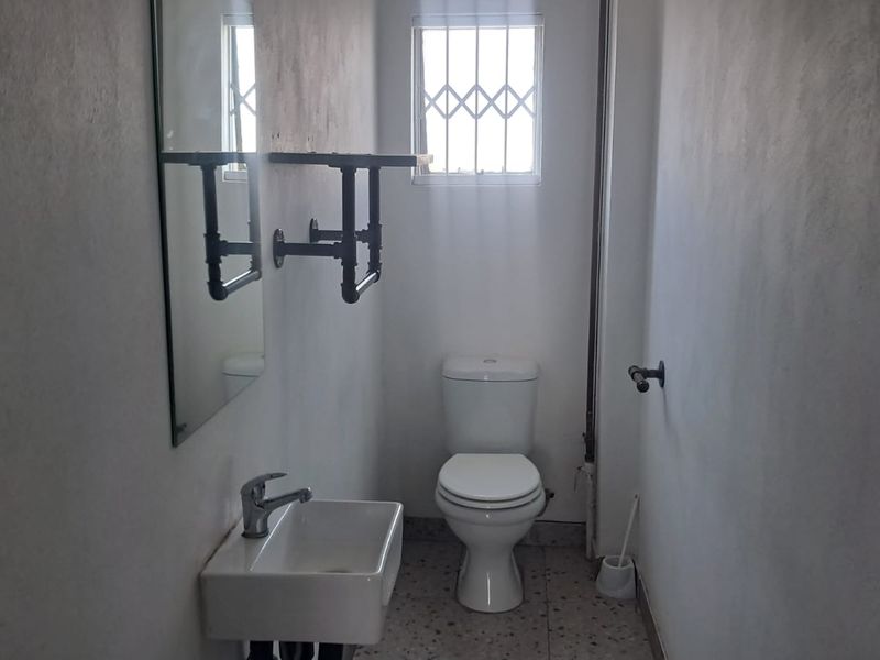 A compact industrial-style bathroom featuring a wall-mounted basin, toilet, and security-barred window, requiring minor cosmetic updates to the wall finishes.