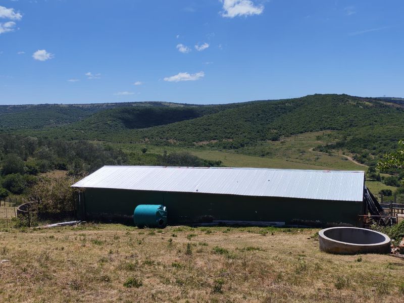 A rear view of a farm building featuring a corrugated metal roof, water storage tank, and concrete trough set against a scenic hilly landscape.