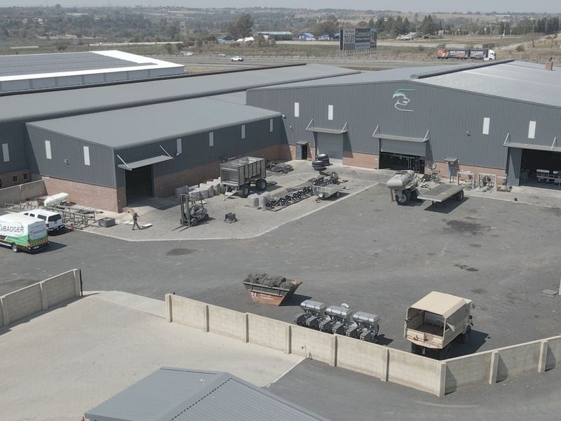 Aerial view of an industrial facility featuring large warehouse structures, metal cladding, expansive asphalt yard space, and perimeter walling.