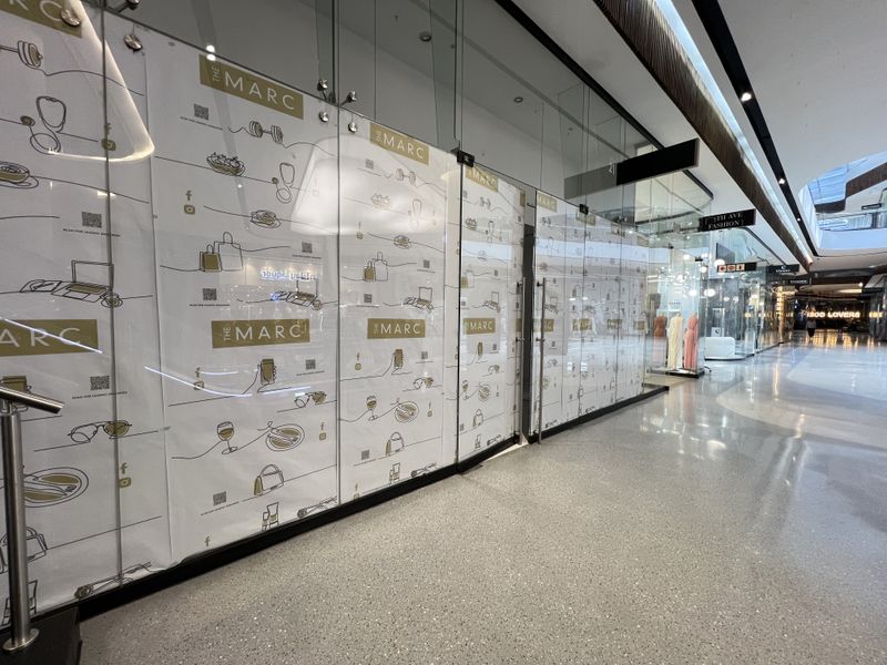 A vacant retail unit within a modern shopping centre, featuring a full-height glass storefront and polished concrete flooring.