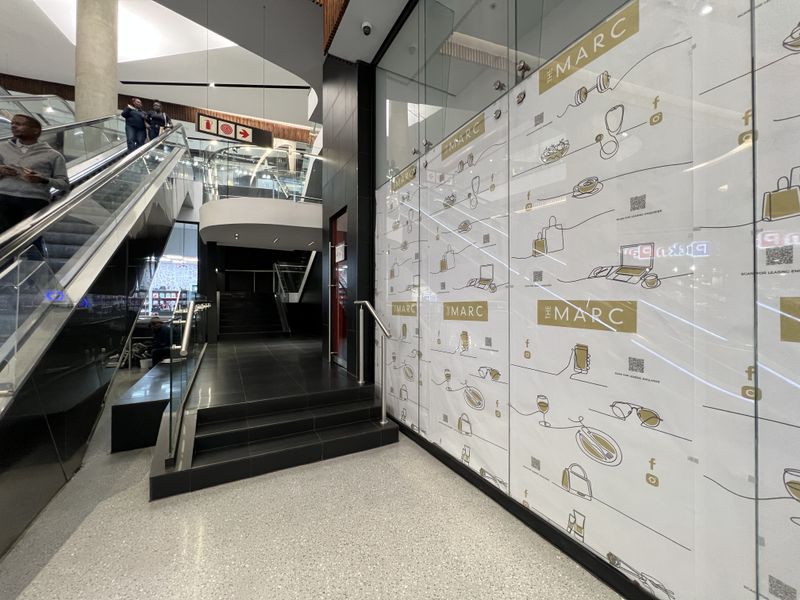 Modern retail entrance area featuring terrazzo flooring, glass storefront display, and an escalator leading to upper levels within a commercial complex.