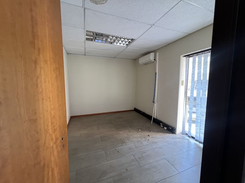 An empty office space featuring tiled flooring, a suspended ceiling with recessed lighting, and a wall-mounted air conditioning unit.