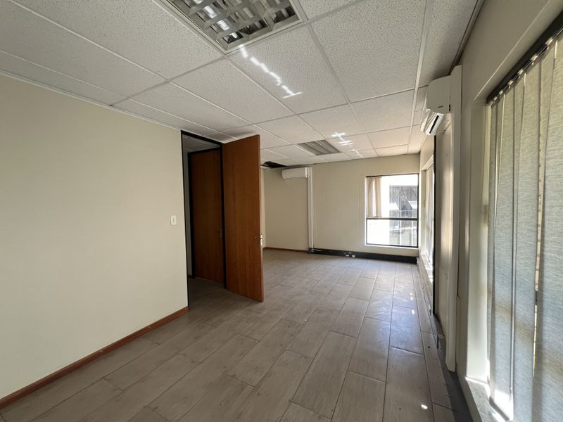 An empty office space featuring tiled flooring, a suspended ceiling with recessed lighting, and wall-mounted air conditioning units.