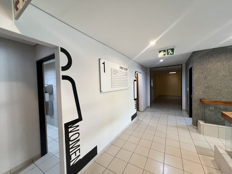 A clean, well-lit office building hallway featuring ceramic floor tiles, clear directional signage, and access to restroom facilities.