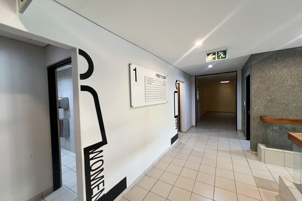 A clean, well-lit office building hallway featuring ceramic floor tiles, clear directional signage, and access to restroom facilities.