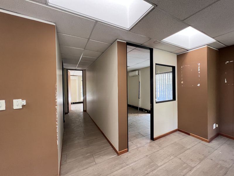 An empty office space featuring tiled flooring, a suspended ceiling with recessed lighting, and an internal glass partition, currently requiring cosmetic wall repairs.