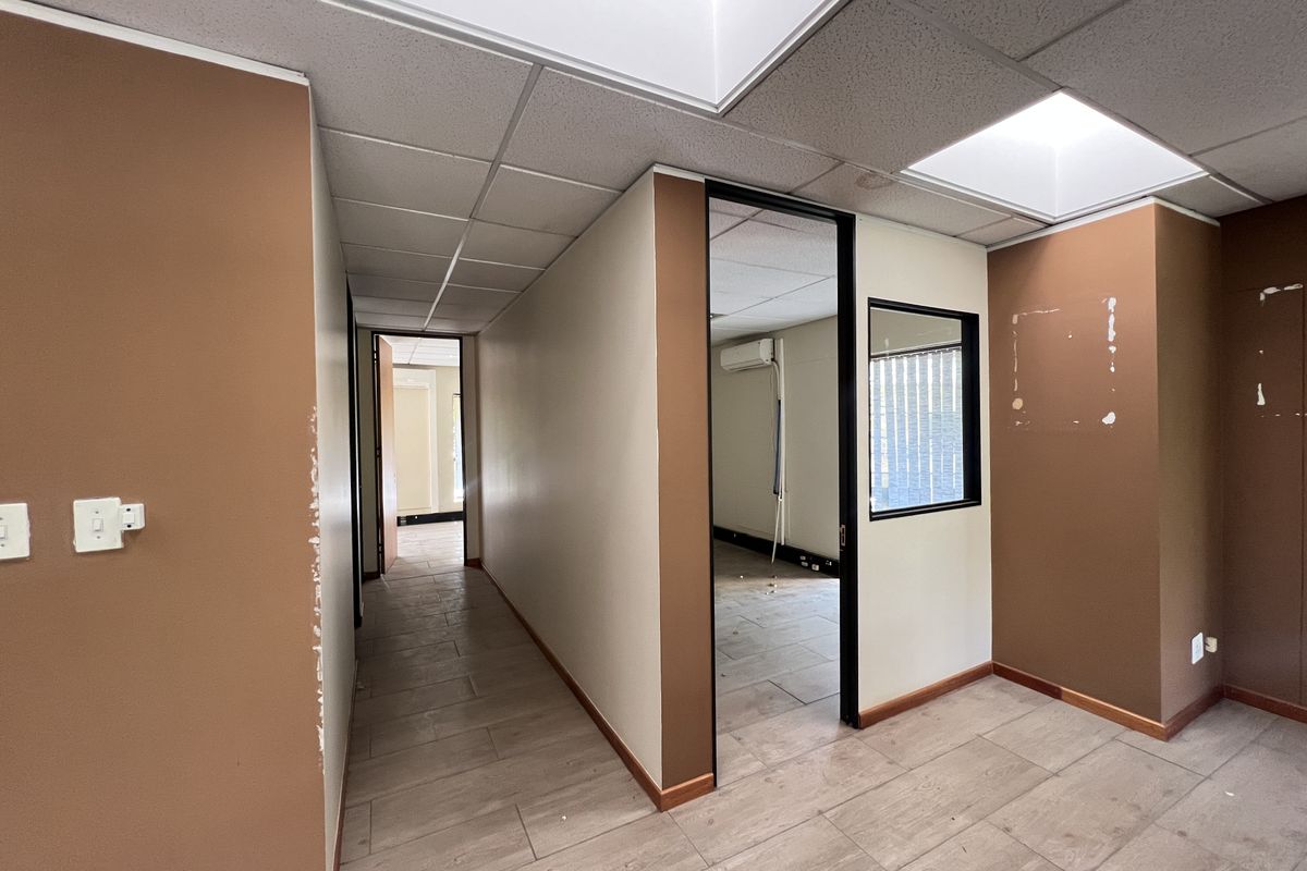 An empty office space featuring tiled flooring, a suspended ceiling with recessed lighting, and an internal glass partition, currently requiring cosmetic wall repairs.