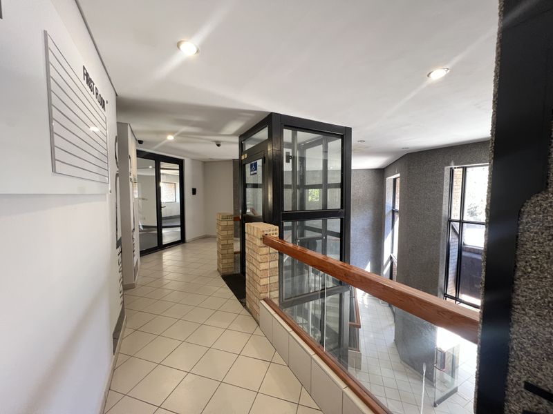 A bright, modern office building hallway featuring a glass elevator, tiled flooring, and a glass balustrade with a wooden handrail.