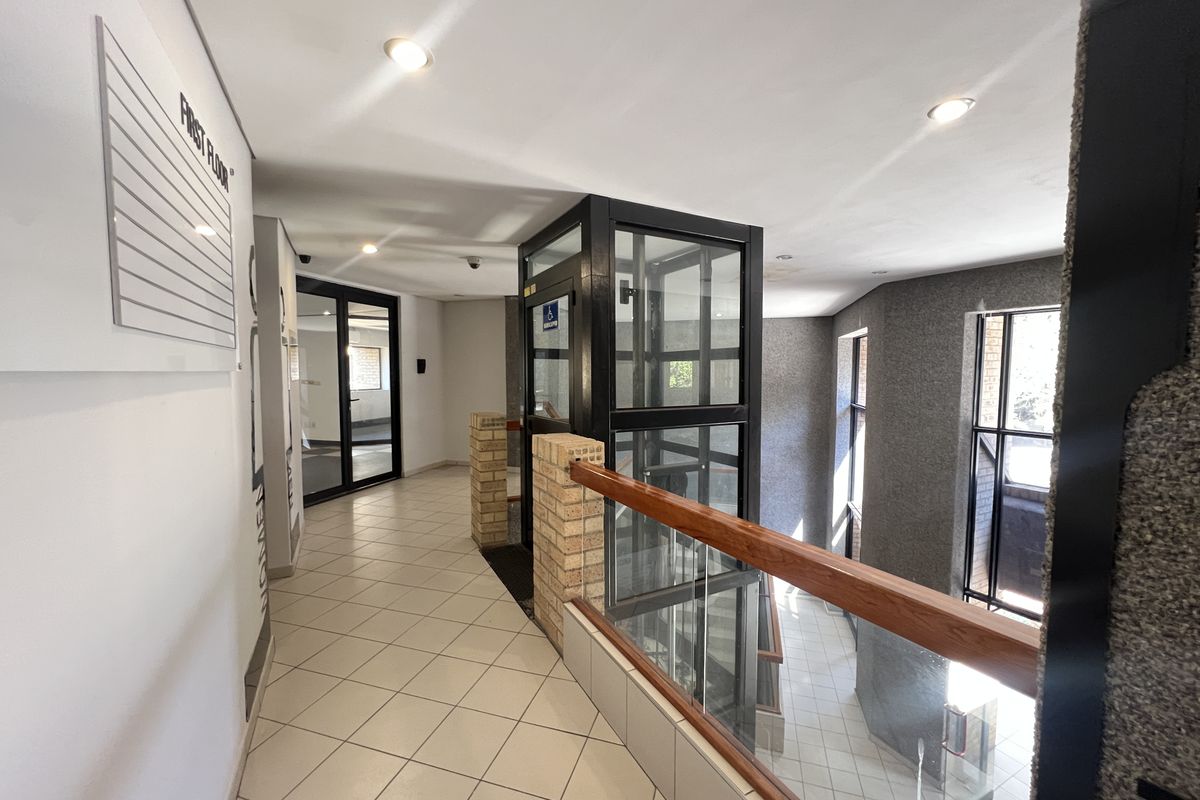 A bright, modern office building hallway featuring a glass elevator, tiled flooring, and a glass balustrade with a wooden handrail.