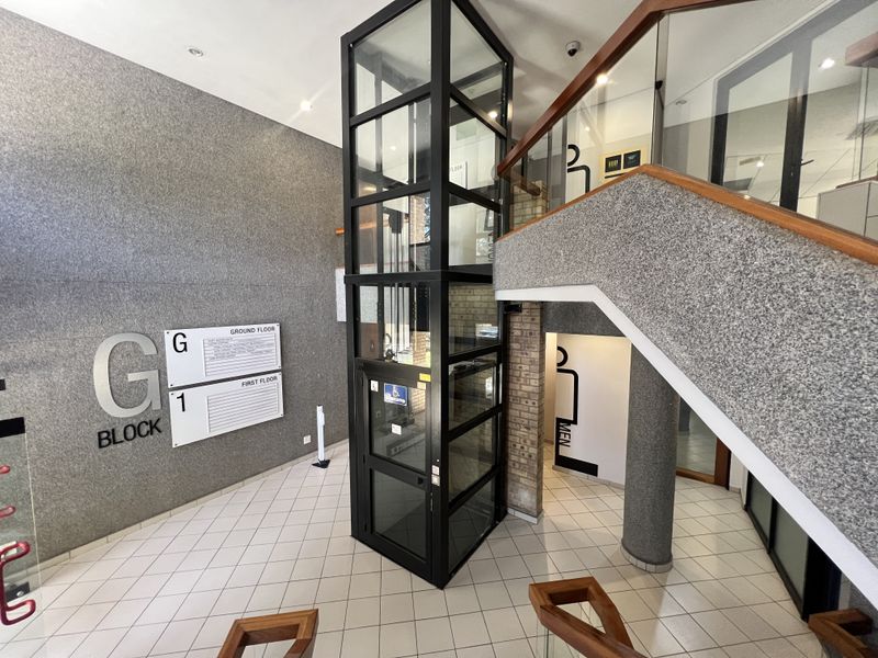 Modern office building lobby featuring a glass elevator, tiled flooring, and a staircase with a glass balustrade and wooden handrail.