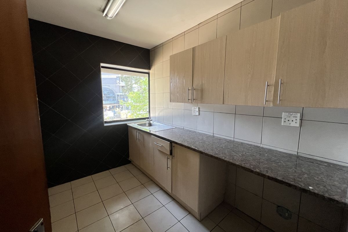 A clean, functional office kitchenette featuring light wood cabinetry, granite countertops, a stainless steel sink, and modern tiled finishes.
