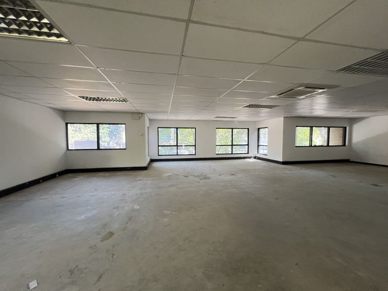 A spacious, empty open-plan office area featuring a suspended ceiling, recessed lighting, integrated air conditioning, and large windows providing natural light.