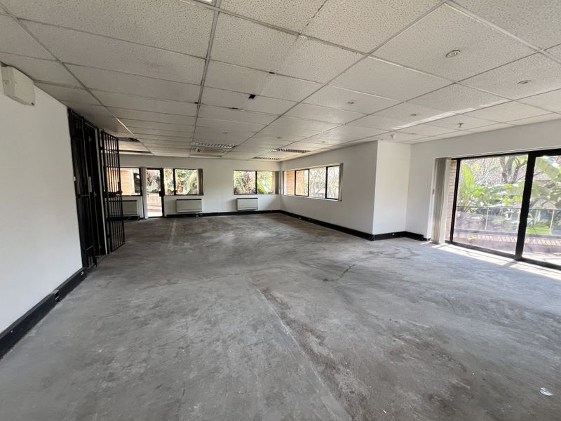 Spacious open-plan office area featuring a suspended ceiling, large windows, sliding glass doors, and security gates, currently in need of flooring and ceiling repairs.