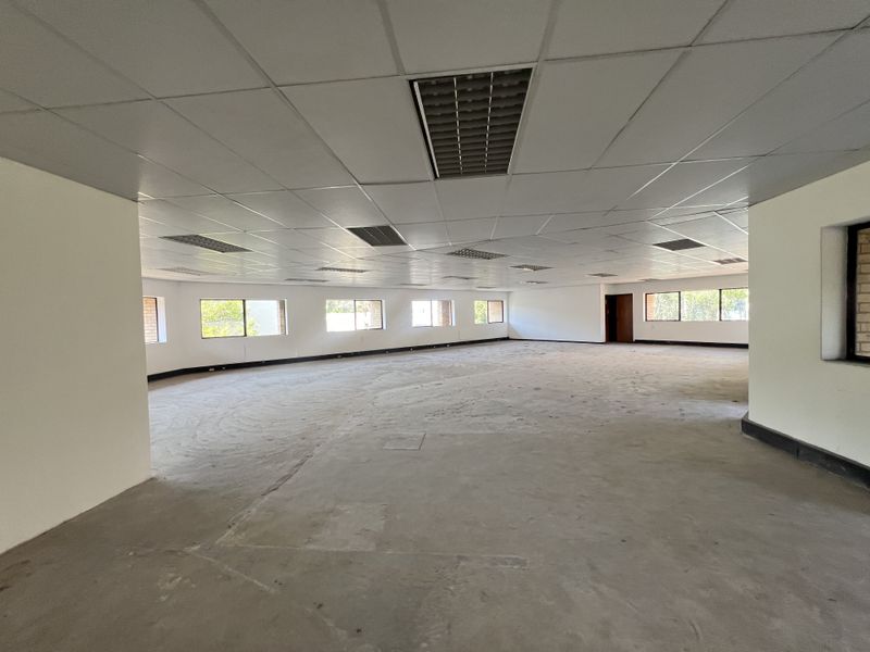 A large, open-plan office space featuring a suspended ceiling with recessed lighting, concrete flooring, and multiple windows providing natural light.