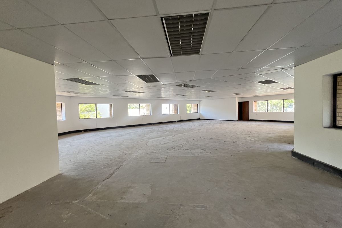 A large, open-plan office space featuring a suspended ceiling with recessed lighting, concrete flooring, and multiple windows providing natural light.
