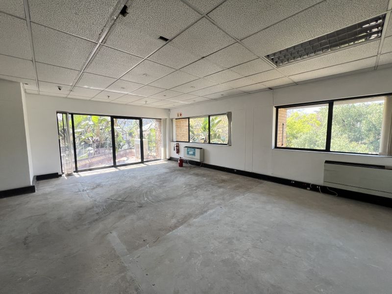 An empty, open-plan office space featuring a suspended ceiling, large windows with natural light, and sliding door access to an outdoor area.
