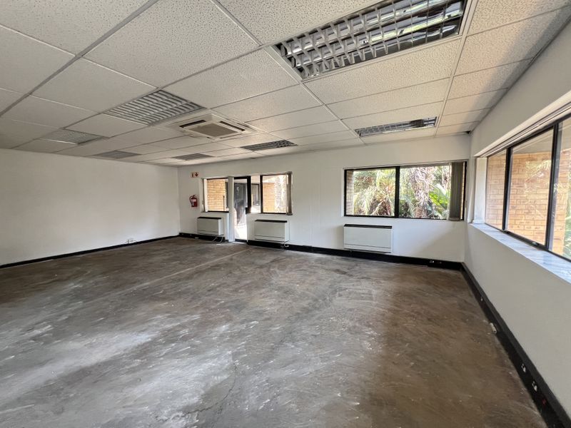 An empty, open-plan office space featuring a suspended ceiling, recessed lighting, air conditioning, and large windows overlooking the exterior.