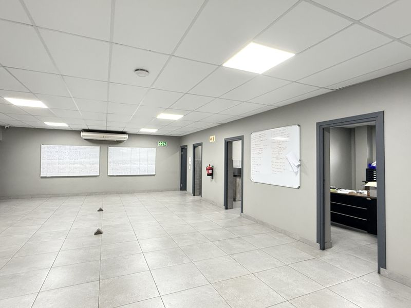 Spacious industrial office area featuring neutral grey walls, ceramic tiled flooring, suspended ceiling with recessed lighting, and wall-mounted air conditioning.