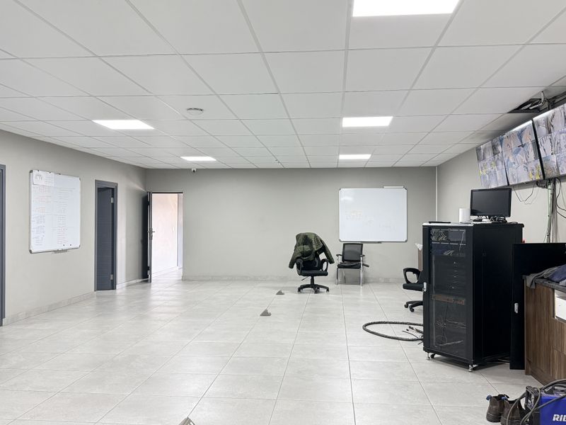 A spacious, well-lit office area featuring tiled flooring, a suspended ceiling, integrated server rack, and a wall-mounted security monitoring system.
