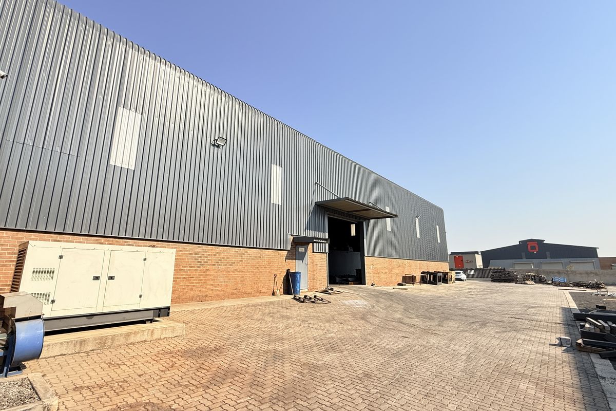 Rear view of an industrial warehouse featuring corrugated metal cladding, brick base, paved yard area, and an external generator unit.
