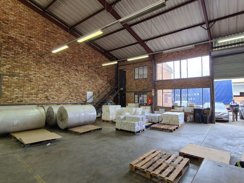 Spacious industrial warehouse featuring exposed brick walls, high steel truss ceilings, concrete flooring, and a large roller shutter door for vehicle access.