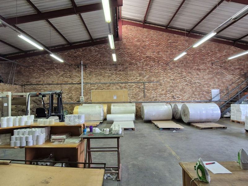Spacious industrial warehouse featuring exposed brick walls, high ceilings with steel trusses, fluorescent lighting, and concrete flooring, suitable for storage or manufacturing operations.