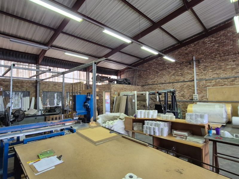Spacious industrial workshop area featuring exposed brick walls, high steel roof trusses, and functional fluorescent lighting, suitable for manufacturing or storage operations.