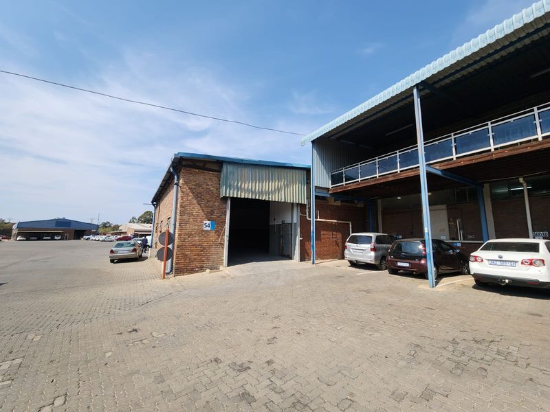 Exterior view of an industrial warehouse unit featuring a large roller shutter door, brick facade, and an elevated mezzanine level with paved parking area.