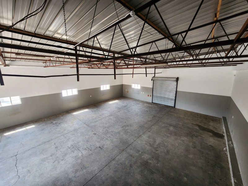 Spacious industrial warehouse unit featuring high exposed steel trusses, concrete flooring, and a large roller shutter door for easy access.