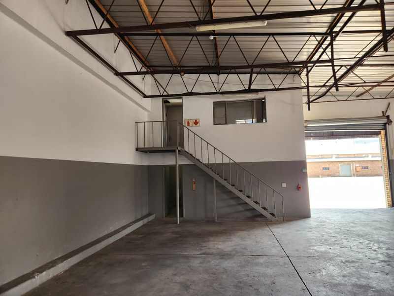 Spacious industrial warehouse unit featuring high ceilings with exposed steel trusses, a mezzanine level with staircase access, and a large roller shutter door.