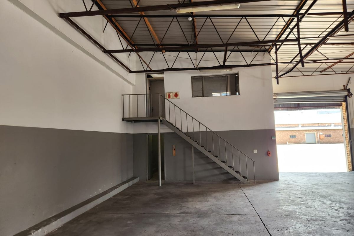 Spacious industrial warehouse unit featuring high ceilings with exposed steel trusses, a mezzanine level with staircase access, and a large roller shutter door.