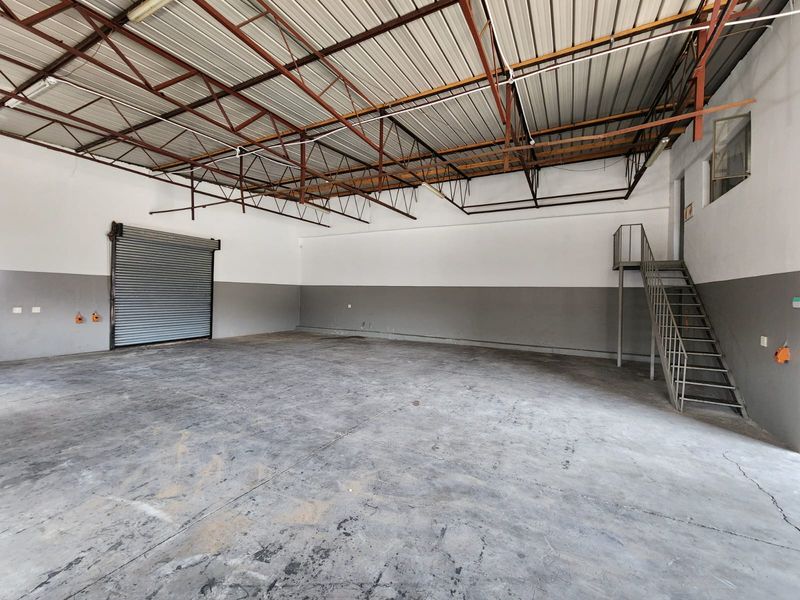 Spacious industrial warehouse unit featuring high ceilings with exposed steel trusses, a roller shutter door, concrete flooring, and a metal staircase to a mezzanine level.