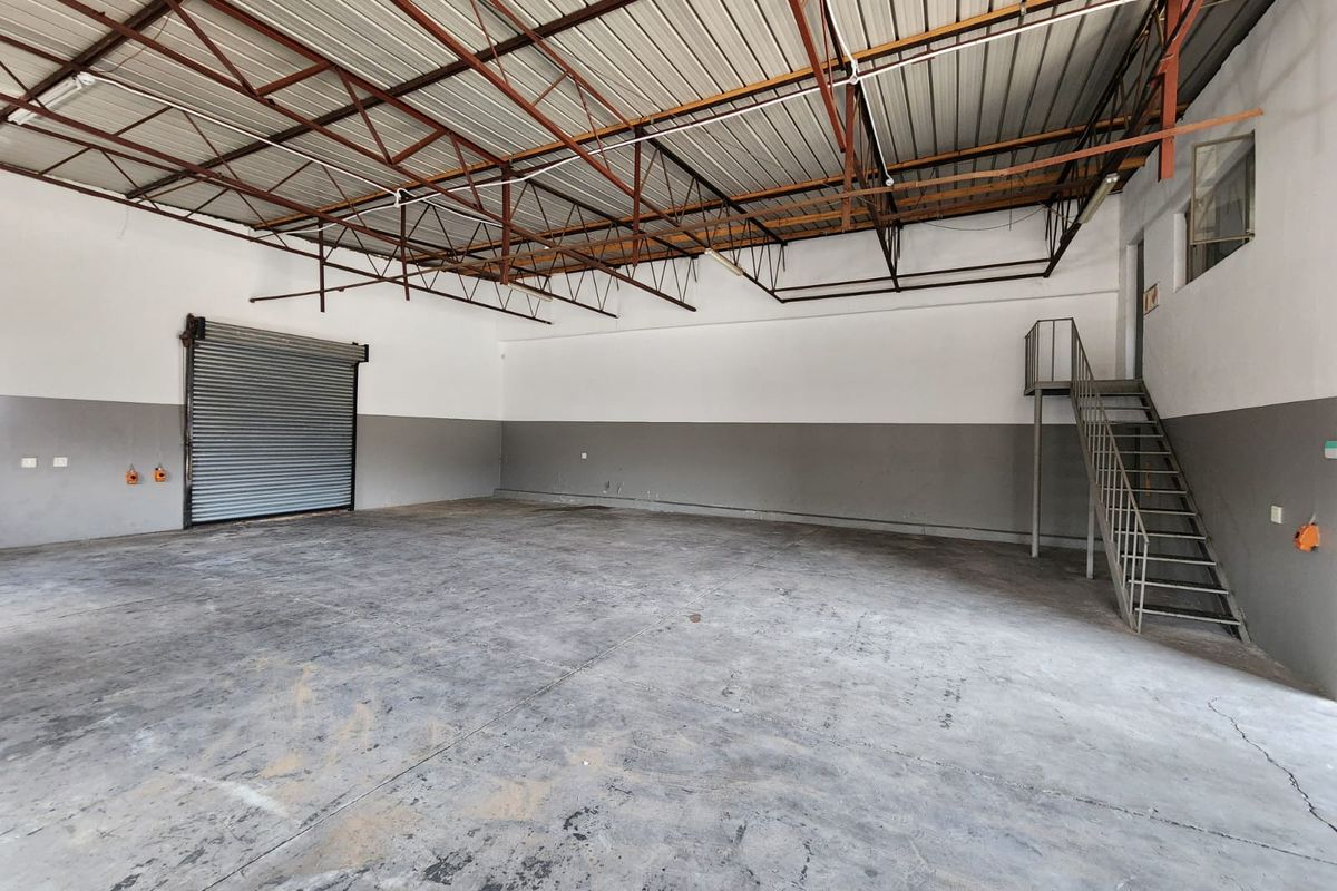 Spacious industrial warehouse unit featuring high ceilings with exposed steel trusses, a roller shutter door, concrete flooring, and a metal staircase to a mezzanine level.