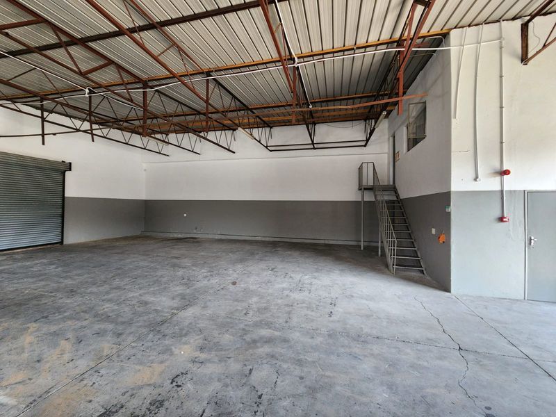 Spacious industrial warehouse unit featuring high exposed steel roof trusses, a large roller shutter door, concrete flooring, and an internal metal staircase.