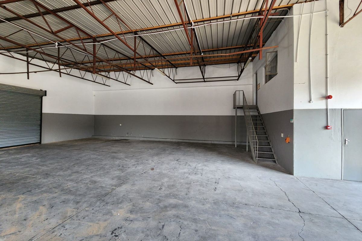 Spacious industrial warehouse unit featuring high exposed steel roof trusses, a large roller shutter door, concrete flooring, and an internal metal staircase.