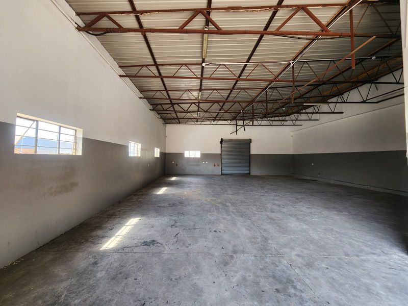 Spacious industrial warehouse unit featuring high exposed steel roof trusses, durable concrete flooring, and a large metal roller shutter door for easy access.
