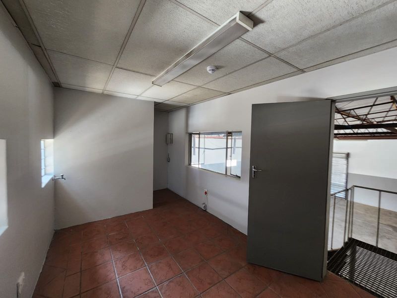 An empty industrial office space featuring tiled flooring, a suspended ceiling with integrated lighting, and direct access to an adjacent mezzanine area.