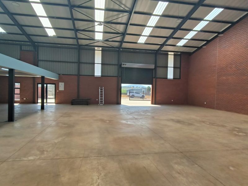 Spacious industrial warehouse featuring high ceilings with exposed steel trusses, translucent roof panels, concrete flooring, and a large roller shutter door for easy access.