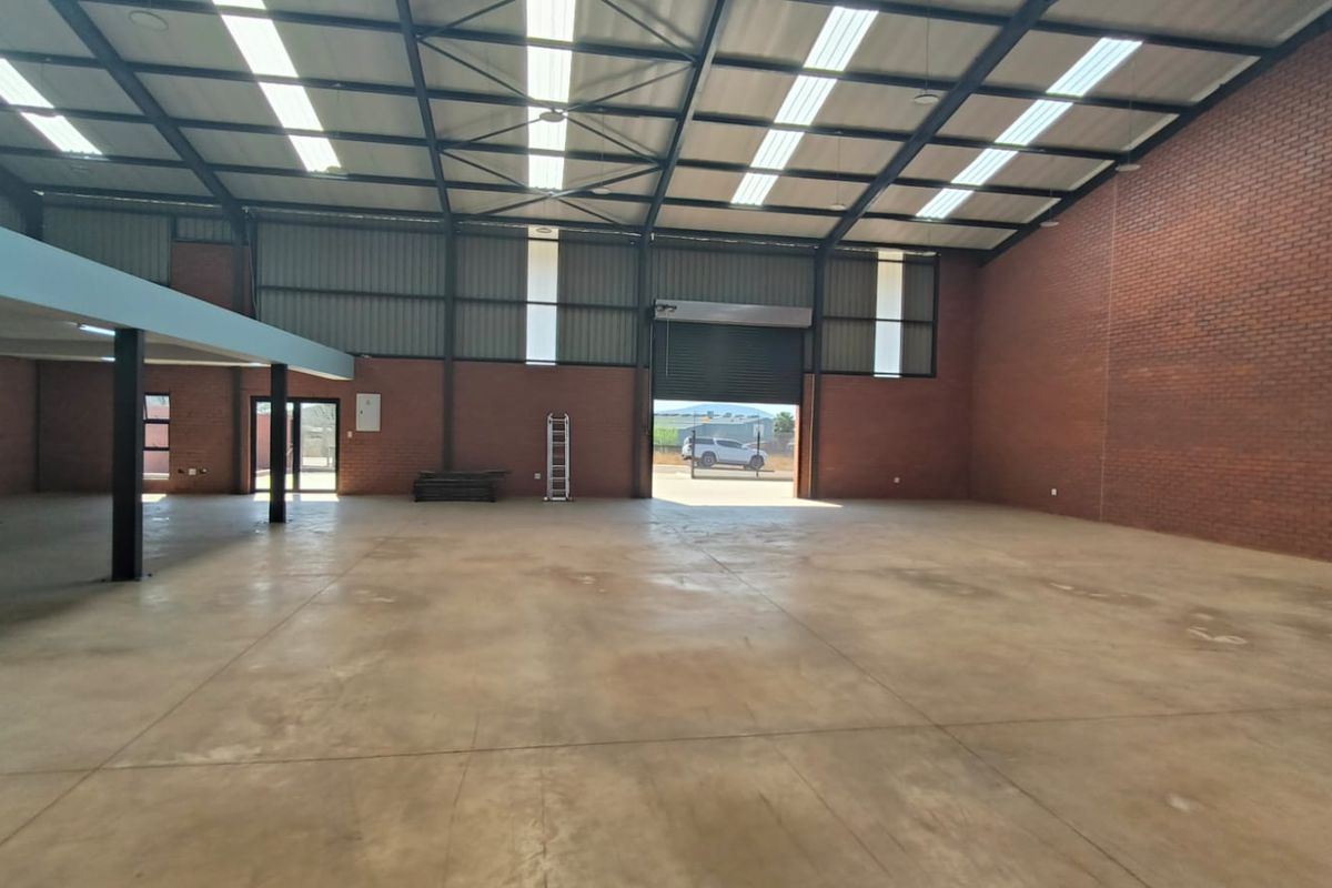 Spacious industrial warehouse featuring high ceilings with exposed steel trusses, translucent roof panels, concrete flooring, and a large roller shutter door for easy access.