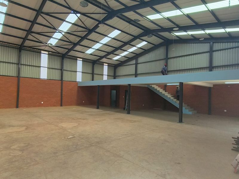Spacious industrial warehouse featuring high ceilings with translucent panels, exposed brick walls, concrete flooring, and a functional mezzanine level with staircase access.