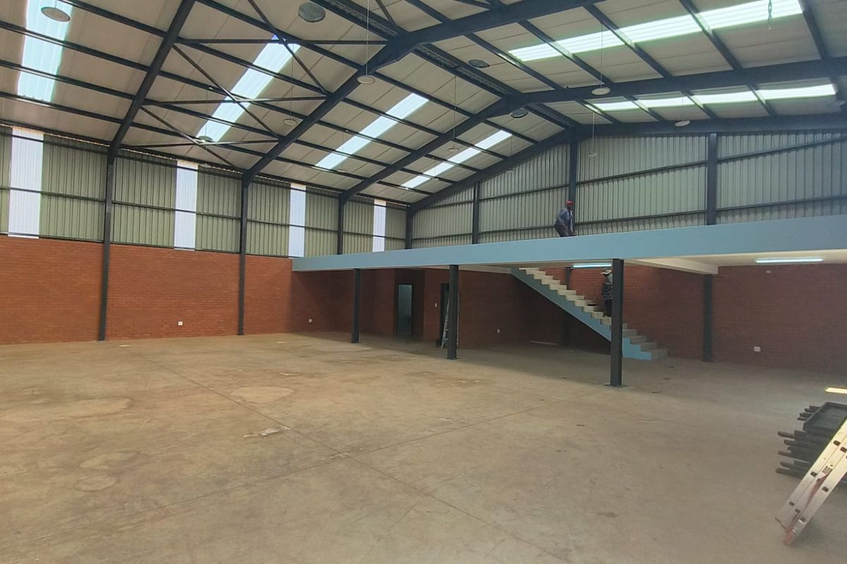 Spacious industrial warehouse featuring high ceilings with translucent panels, exposed brick walls, concrete flooring, and a functional mezzanine level with staircase access.