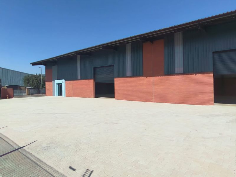 Industrial warehouse exterior featuring a large paved yard, multiple roller shutter doors, and a combination of brick and metal cladding construction.
