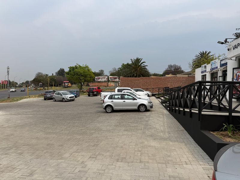 Exterior view of a commercial property featuring a paved parking area, brick boundary wall, and modern black metal railings leading to the building entrance.