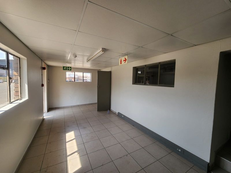 An empty industrial hallway featuring tiled flooring, a suspended ceiling with fluorescent lighting, and clear fire safety signage.