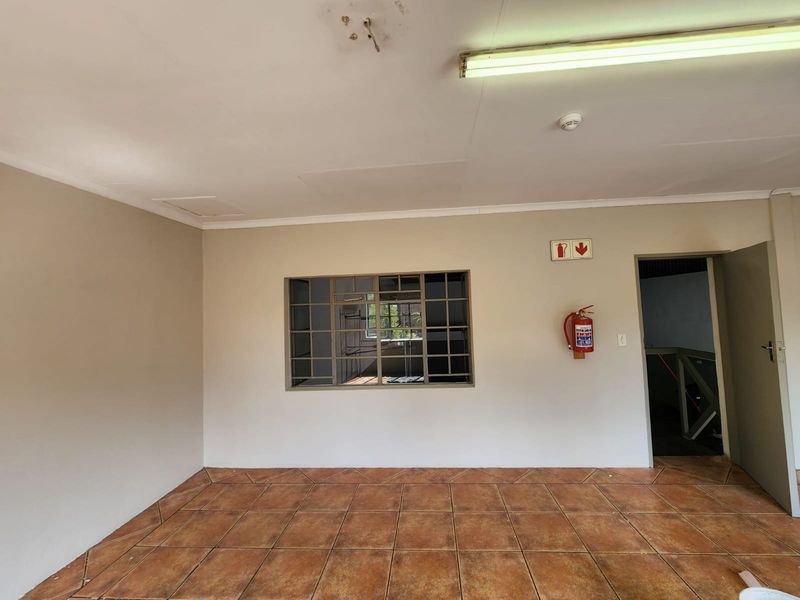 An empty industrial office space featuring tiled flooring, a steel-framed window, and essential fire safety equipment including an extinguisher and smoke detector.